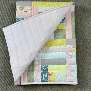 May include: A handmade quilt with a patchwork design. The quilt features a variety of fabrics in pastel colors, including pink, green, yellow, and gray, with floral and dotted patterns. The quilt's underside is a solid light pink, and the edges are neatly bound.