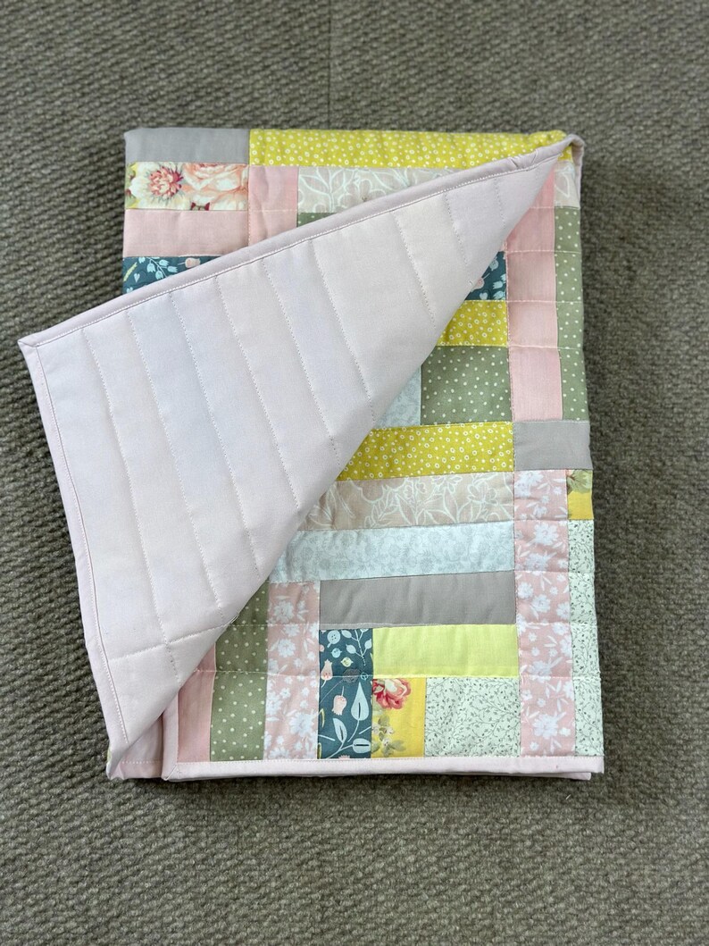 May include: A handmade quilt with a patchwork design. The quilt features a variety of fabrics in pastel colors, including pink, green, yellow, and gray, with floral and dotted patterns. The quilt's underside is a solid light pink, and the edges are neatly bound.