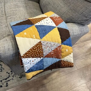 Puede incluir: Un edred&oacute;n patchwork hecho a mano con un dise&ntilde;o geom&eacute;trico. El edred&oacute;n presenta tri&aacute;ngulos en varios colores y patrones, incluyendo azul, amarillo, marr&oacute;n y blanco. El edred&oacute;n est&aacute; doblado sobre el brazo de un sof&aacute; gris, mostrando sus texturas y colores.
