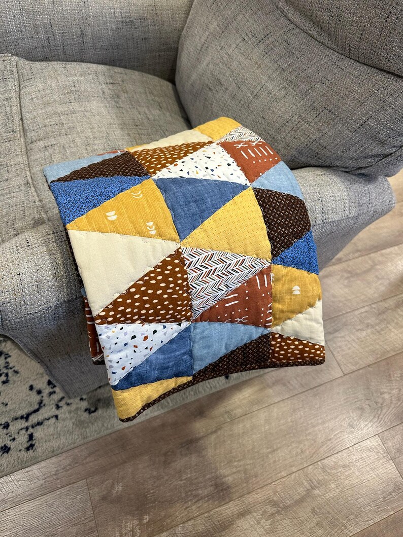Puede incluir: Un edred&oacute;n patchwork hecho a mano con un dise&ntilde;o geom&eacute;trico. El edred&oacute;n presenta tri&aacute;ngulos en varios colores y patrones, incluyendo azul, amarillo, marr&oacute;n y blanco. El edred&oacute;n est&aacute; doblado sobre el brazo de un sof&aacute; gris, mostrando sus texturas y colores.