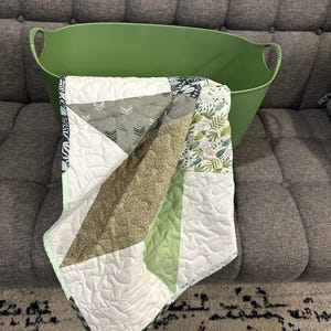 May include: A green oval-shaped storage bin holds a patchwork quilt. The quilt features geometric patterns in white, green, gray, and olive green. The quilt has a white backing and a dark green border.