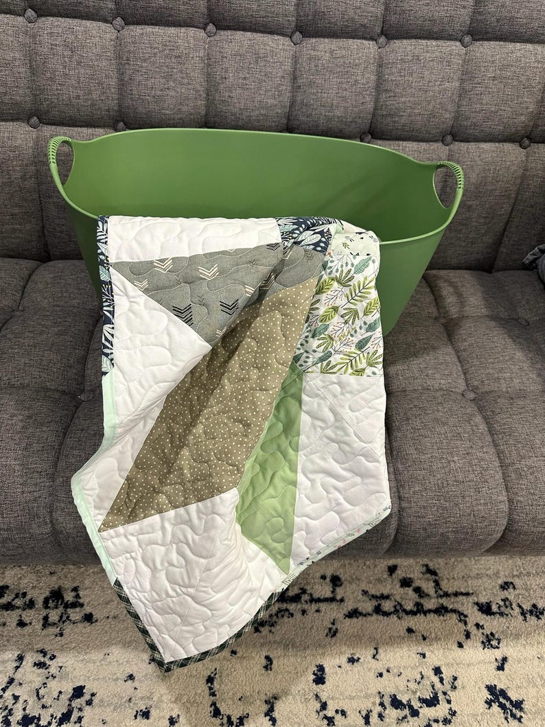 May include: A green oval-shaped storage bin holds a patchwork quilt. The quilt features geometric patterns in white, green, gray, and olive green. The quilt has a white backing and a dark green border.