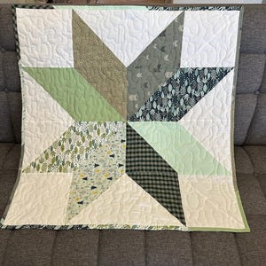 May include: A handmade quilt featuring a starburst design. The quilt showcases various fabrics in shades of green, white, and gray, with a plaid and leaf pattern. The quilt is approximately 40 inches square.