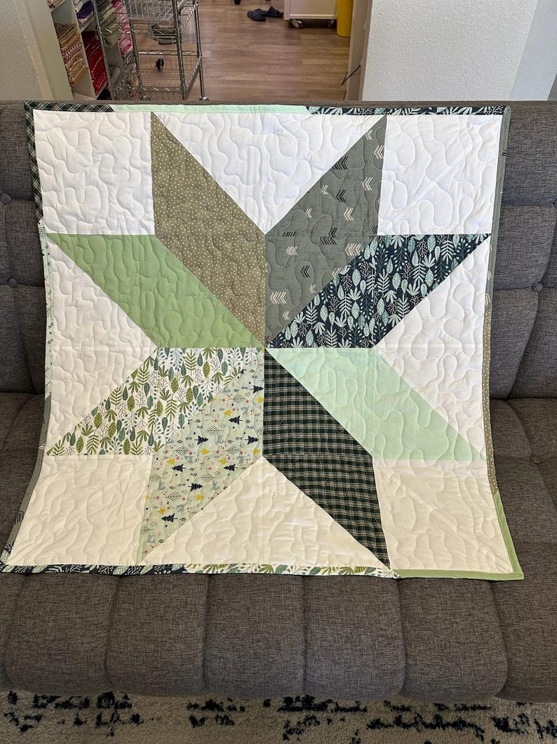 May include: A handmade quilt featuring a starburst design. The quilt showcases various fabrics in shades of green, white, and gray, with a plaid and leaf pattern. The quilt is approximately 40 inches square.