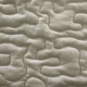 May include: Close-up of a white quilted fabric with a cloud-like pattern. The stitching follows the contours of the shapes, creating a textured surface. The fabric appears to be a soft, light material.
