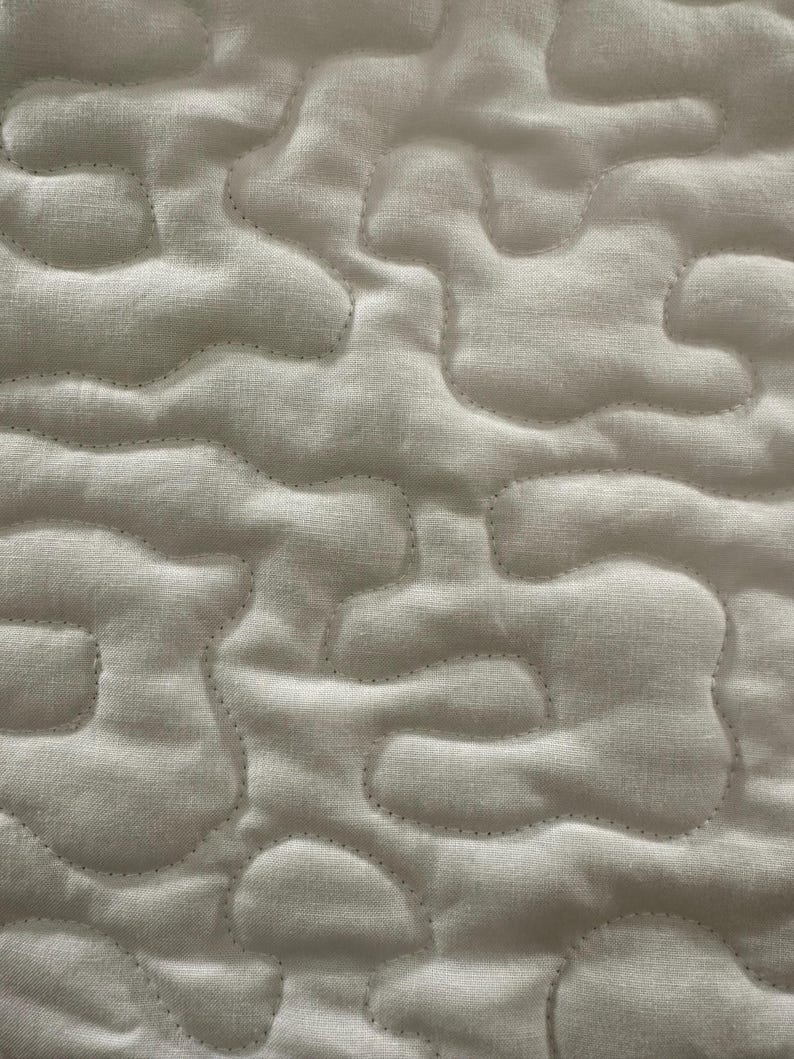 May include: Close-up of a white quilted fabric with a cloud-like pattern. The stitching follows the contours of the shapes, creating a textured surface. The fabric appears to be a soft, light material.