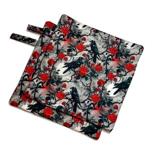 以下が含まれることがあります： 黒いカラス、赤いバラ、白いドクロの繰り返し模様がグレーの背景にプリントされた、正方形の布製鍋つかみ3枚。各鍋つかみには、吊り下げ用の黒い布ループが付いています。