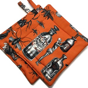 May include: Two orange fabric pot holders with a Halloween theme. The pot holders feature a repeating pattern of bottles, insects, and the text "Dark Magic Potion" and "Octopus Ink".