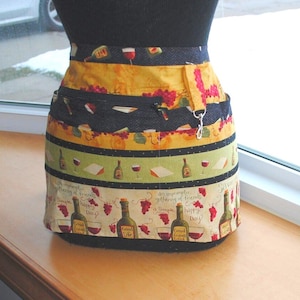 Puede incluir: Un delantal estampado en amarillo, verde y negro con un diseño de botella de vino tinto y blanco. El delantal tiene varios bolsillos y una correa con un llavero. El texto en el delantal dice "una reunión improvisada de gente amigable", "feliz día", y "buena vida".