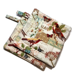 Puede incluir: Dos agarraderas de olla acolchadas de color crema con un diseño floral y de pájaros. La tela presenta un patrón de inspiración vintage con pájaros, mariposas, flores y notas musicales. El texto "LAIS" es visible en la tela.