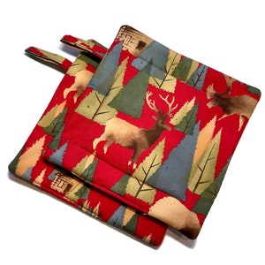 May include: Two red quilted pot holders with a forest scene featuring brown deer, green and blue trees, and small cabins. Each pot holder has a fabric loop for hanging.