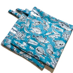 Può includere: Due presine blu turchese con una stampa di mercato del pesce bianca e nera. La stampa presenta vari tipi di pesce, frutti di mare e le parole "Mercato del pesce", "Frutti di mare" e "Pesce fresco".