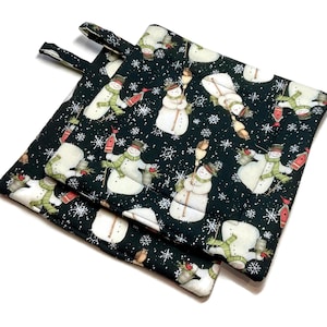 May include: Two square, quilted pot holders with a black background and a repeating pattern of snowmen, snowflakes, and birds. The snowmen wear hats, scarves, and hold birdhouses. Each pot holder has a small fabric loop for hanging.