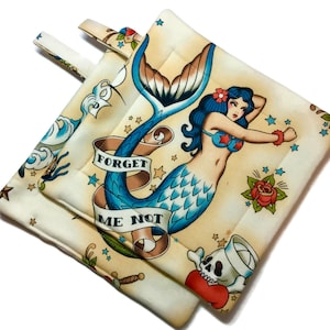 May include: A pair of beige pot holders with a nautical theme. The pot holders feature a mermaid with blue and green scales, a red rose, and the text "Forget Me Not".
