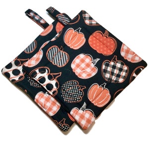May include: A pair of black pot holders with a pumpkin print. The pumpkins are in various patterns, including polka dots, stripes, and plaid. The pot holders have a loop handle.