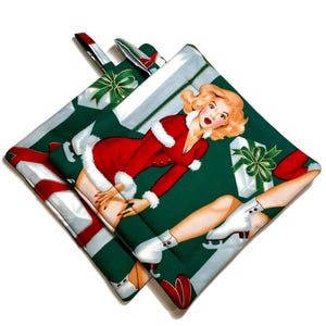 May include: A pair of green and white pot holders with a Christmas theme. The pot holders feature a woman in a red and white Santa suit with a white fur trim, holding a gift box. The woman is wearing ice skates and is standing on a green background with white gift boxes.