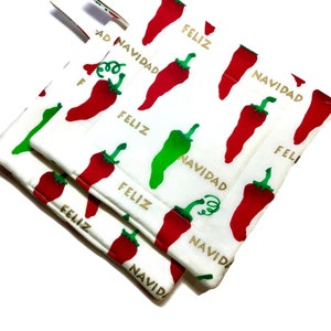 May include: Two white fabric pot holders with a red and green chili pepper pattern. The text "Feliz Navidad" is repeated on the pot holders in gold lettering.