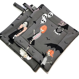 May include: Two gray fabric pot holders with a Halloween theme. The pot holders feature a black and white illustration of girls with bats and umbrellas. One pot holder has a hanging loop.