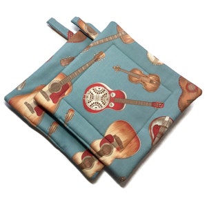 May include: Two square pot holders with a light blue background and a repeating pattern of guitars, violins, and other stringed instruments. Each pot holder has a loop for hanging.