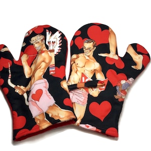Peut inclure: Une paire de maniques de four rouge et noir sur le thème de la Saint-Valentin. Les maniques présentent des images de deux hommes, l'un avec des ailes et une flèche, l'autre tenant un livre. Les maniques sont décorées de cœurs rouges.