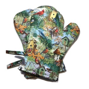 May include: A pair of oven mitts with a colorful bird and birdhouse pattern. The mitts feature a vibrant design of various birds, birdhouses, and foliage in shades of green, blue, red, and brown. They have a loop for hanging and are made of a durable fabric.