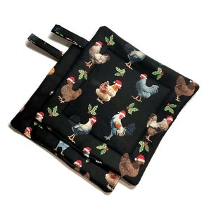 Puede incluir: Un par de agarraderas de olla acolchadas negras con un patrón de pollo navideño. Los pollos llevan gorros de Papá Noel y hay ramas de acebo en todo el diseño.