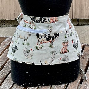 May include: A cream-colored waist apron featuring a farm animal print, including cows, pigs, chickens, and sheep. The apron has a white zipper pocket and ties at the waist. The fabric is patterned with green grass and floral accents.