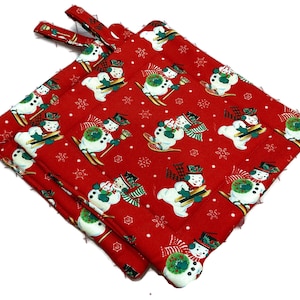 Peut inclure: Une paire de maniques carrées matelassées présentant un fond rouge vif avec un motif répétitif de bonhommes de neige blancs portant des chapeaux et des écharpes, agrémenté de petits flocons de neige blancs et de couronnes vertes.