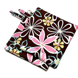 Könnte beinhalten: Braune und rosa Ofenhandschuhe mit Blumendruck. Die Handschuhe zeigen ein sich wiederholendes Muster aus großen Blumen in Rosa, Weiß und Gelb auf braunem Hintergrund. Jeder Handschuh hat eine Schlaufe zum Aufhängen.