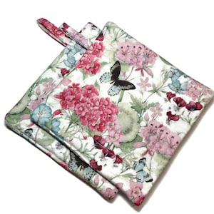 May include: A pair of white cotton pot holders with a floral print featuring pink, blue, and green flowers and butterflies. The pot holders have a loop handle.
