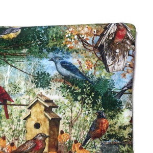 May include: A colorful fabric with a bird and birdhouse theme. The fabric features a variety of birds, including a blue jay, a cardinal, and a robin, perched on branches and in birdhouses. The background is a watercolor-style depiction of trees and leaves.