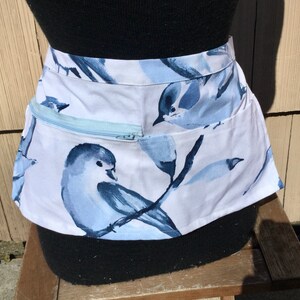 May include: A white apron with a blue bird print and a zippered pocket. The apron is tied at the waist and has a blue and white design.