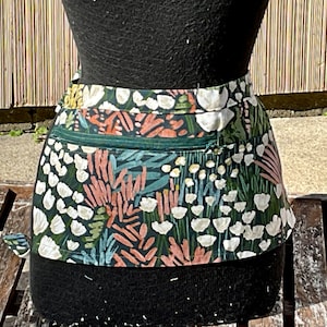 Może przedstawiać: Zielony, biały i różowy fartuch z kwiatowym nadrukiem i kieszenią zapinaną na zamek błyskawiczny. Fartuch jest wykonany z bawełnianej tkaniny i ma czarny pas.