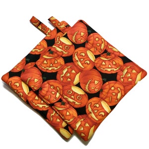 Puede incluir: Un par de agarraderas de olla negras con temática de Halloween. Las agarraderas de olla están cubiertas con un patrón de caras de calabaza naranja.