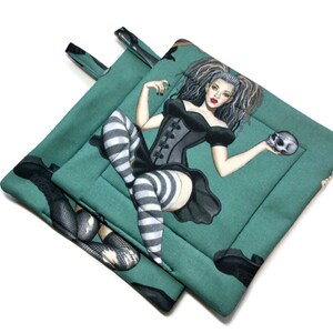 May include: A pair of teal green pot holders with a black and white illustration of a woman in a black corset and striped stockings holding a skull.