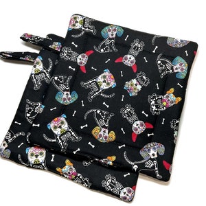 Op de afbeelding: Twee zwarte ovenwanten met een kleurrijk Day of the Dead hondenpatroon. De honden zijn skeletten met bloemen en andere versieringen op hun schedels.