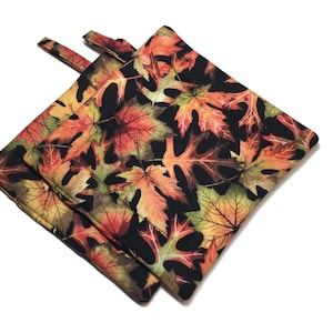 May include: Two square pot holders with a fall leaf pattern. The fabric features red, orange, yellow, and green leaves on a black background. Each pot holder has a small loop for hanging.
