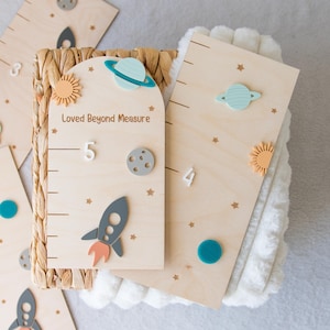 May include: Wooden growth charts with space-themed decorations. The charts feature rockets, planets, suns, and stars in various colors. One chart reads "Loved Beyond Measure". The charts are designed to measure height in inches.