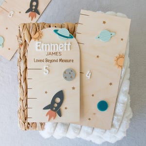 May include: Wooden height chart with space-themed decorations. The chart features a rocket, planets, and stars, with the name "Emmett" and the phrase "Loved Beyond Measure". The chart is designed to measure height in centimetres.