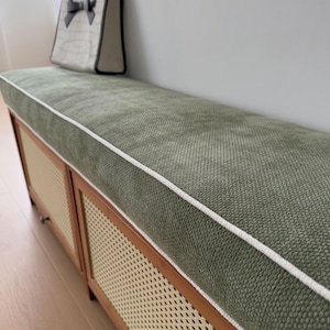 Puede incluir: Banco tapizado verde con ribete blanco y marco de madera. El banco tiene un diseño de caña tejida en la parte delantera y un cojín verde a juego. Una bolsa tote beige con un lazo negro está encima.