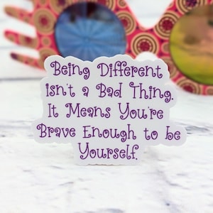 May include: White sticker with purple text that reads "Being Different isn't a Bad Thing. It Means You're Brave Enough to be Yourself." The sticker is sitting on a white surface with a pair of pink and gold glasses in the background.