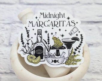 May include: White sticker with black text that reads "Midnight Margaritas" with a black and white illustration of a bottle of tequila, a frog sitting on a mushroom, a salt shaker, a lime wedge, and a margarita glass. The sticker is on a white marble mortar and pestle.