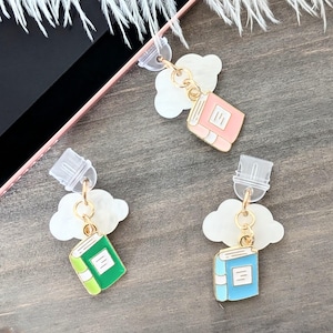 May include: Three phone charms featuring a book design. Each charm is attached to a clear plastic phone grip with a white cloud-shaped charm. The books are pink, green, and blue.