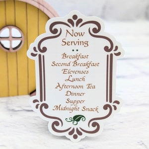 May include: A white sign with brown decorative borders and green accents. The sign reads "Now Serving" and lists various meal times: Breakfast, Second Breakfast, Elevenses, Lunch, Afternoon Tea, Dinner, Supper, and Midnight Snack.