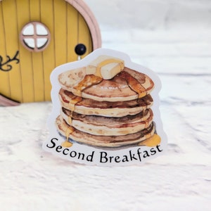 Peut inclure: Un autocollant blanc avec une illustration aquarelle d'une pile de crêpes avec du beurre et du sirop. Le texte "Second Breakfast" est imprimé sous les crêpes.