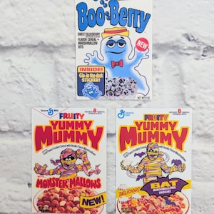 Pode incluir: Três caixas de cereais vintage. A primeira caixa é branca com um personagem de monstro Boo-Berry azul e roxo. O texto na caixa diz "Boo-Berry Sweet Blueberry Flavor Cereal + Marshmallow Bits" e "New! Inside! Glo-in-the-dark Sticker!" A segunda caixa é vermelha e branca com um personagem de múmia e o texto "Fruity Yummy Mummy Fruit Flavor Frosted Cereal with Vanilla Flavor Marshmallows with... Monster Mallows! New!" A terceira caixa é vermelha e branca com um personagem de múmia e o texto "Fruity Yummy Mummy Fruit Flavor Frosted Cereal with Vanilla Flavor Marshmallows Delicious! Bat Marshmallows Inside!"