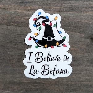 May include: A white sticker with a black witch hat decorated with colorful Christmas lights. The text "I Believe in La Befana" is written in black script below the hat.