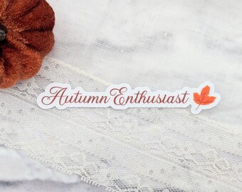 以下が含まれることがあります： 筆記体で「Autumn Enthusiast」と書かれたステッカー。オレンジ色の葉のイラストが添えられています。白いレースの表面に貼られており、左上に小さな茶色のベルベットのカボチャがあります。