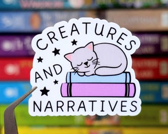 May include: A white sticker with the text "CREATURES AND NARRATIVES" and a sleeping cat illustration. The cat is light purple and rests on a stack of books. The books are pink and blue. Black stars are scattered around the text.