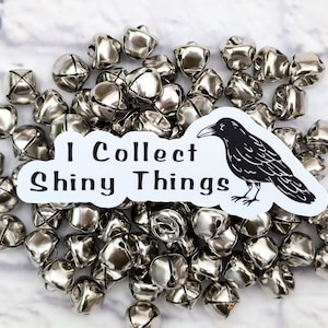 Puede incluir: Una colección de pequeñas campanas plateadas brillantes dispersas sobre una superficie. Una pegatina blanca con el texto "I Collect Shiny Things" y una ilustración de cuervo negro se coloca entre las campanas. Las campanas tienen forma cuadrada con un diseño de cruz.